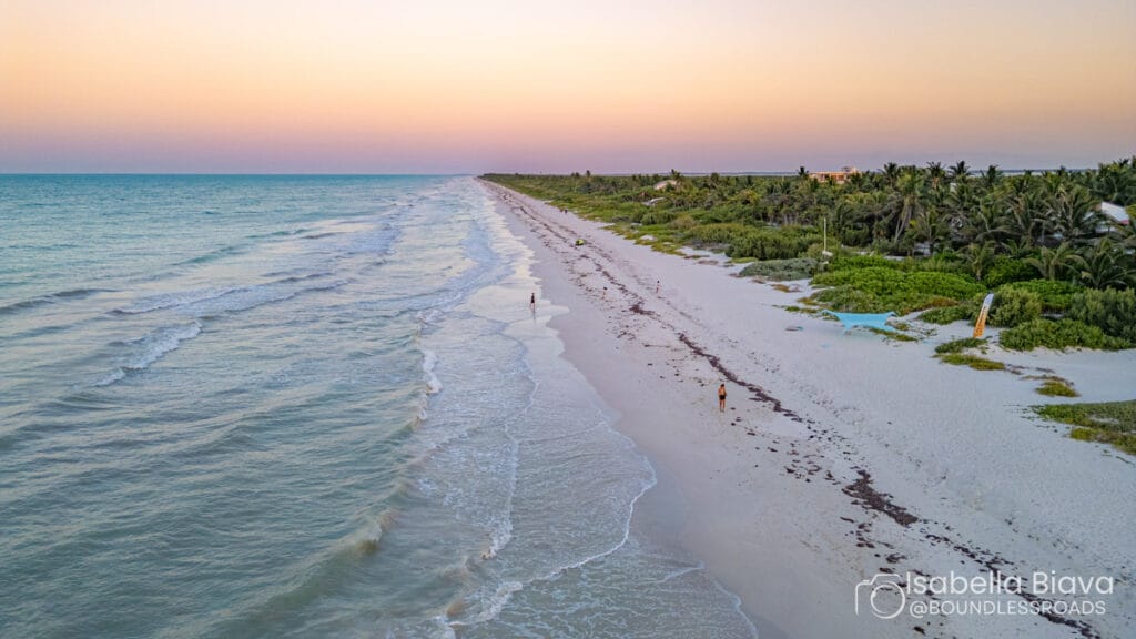 A tranquil beach scene at sunrise or sunset, featuring a long stretch of shoreline, with a few people walking along the water's edge.