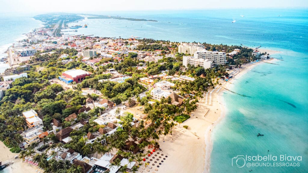 Aerial view of Isla Mujeres, featuring sandy beaches, turquoise waters, lush greenery, and coastal buildings, showcasing a vibrant tropical island atmosphere.