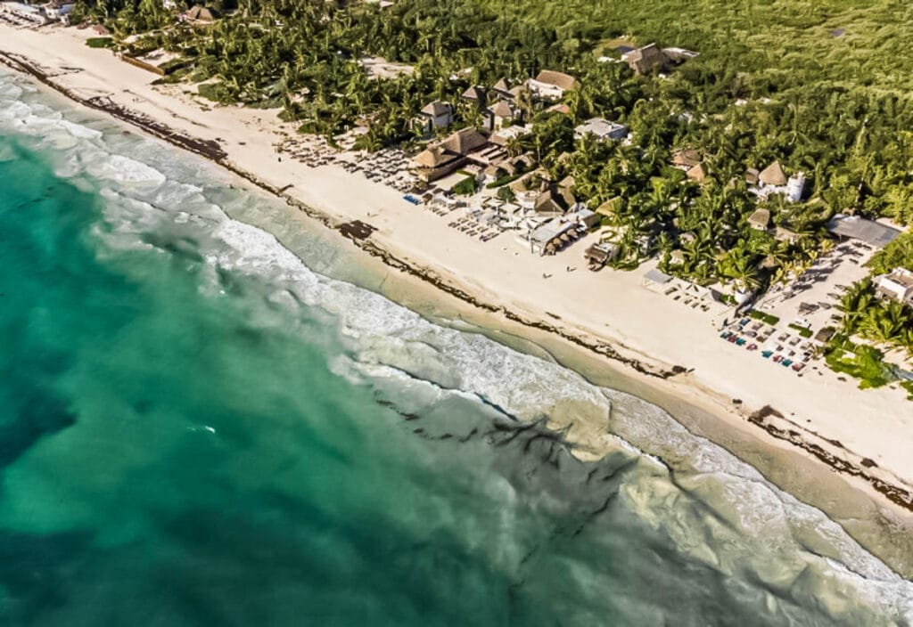 Aerial view of a scenic beach with turquoise waters, white sand, beachfront buildings, and green foliage. People relax under umbrellas along the shore.