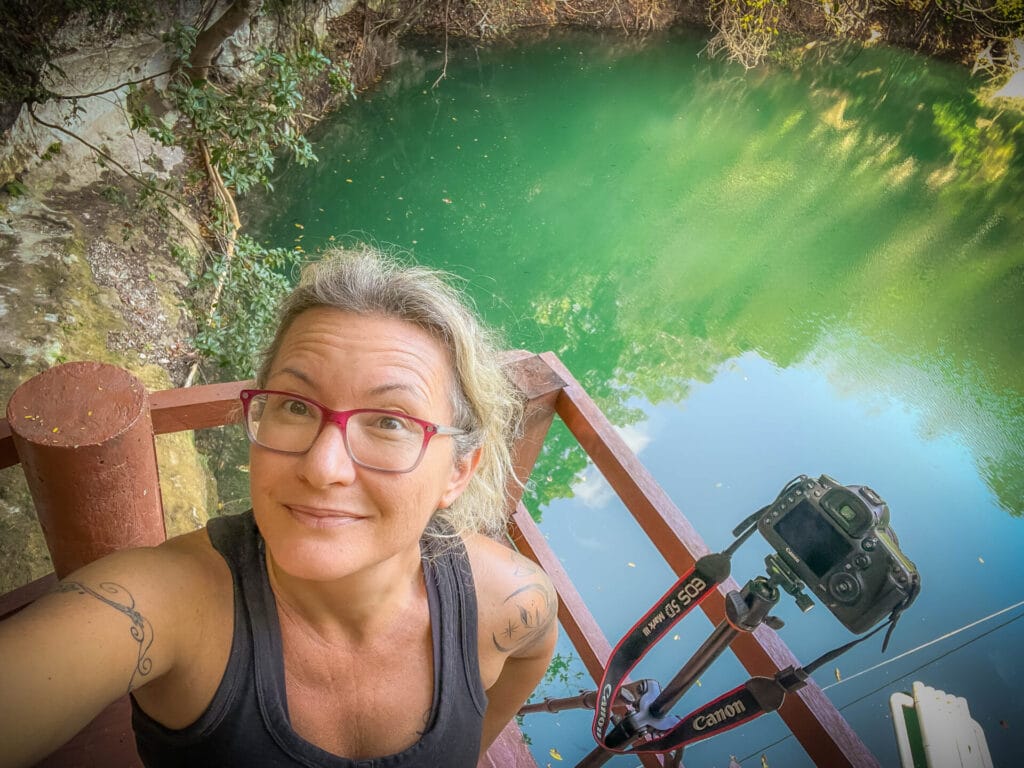 A person with glasses and tattoos stands near a serene green pond, holding a Canon camera on a wooden platform amidst lush surroundings.
