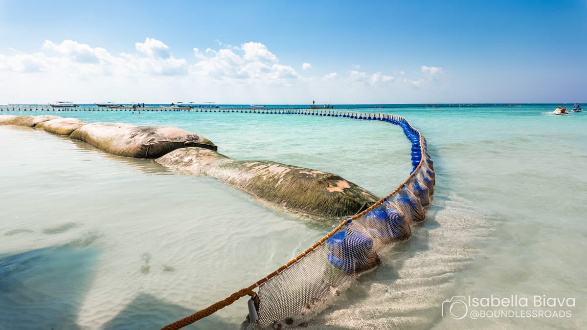 A picturesque beach features clear turquoise waters, a sand barrier, and scattered people in the distance, under a bright, sunny sky.