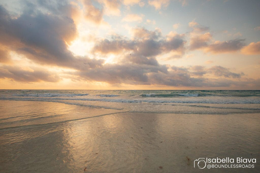 Sunset over calm ocean waves at a sandy beach, with dramatic clouds reflecting warm light. Tranquil and scenic seascape view.